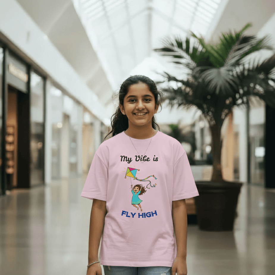 A young girl smiling in a shopping mall wearing a pink "Fly High" Kids Pongal T-shirt featuring a playful kite design.